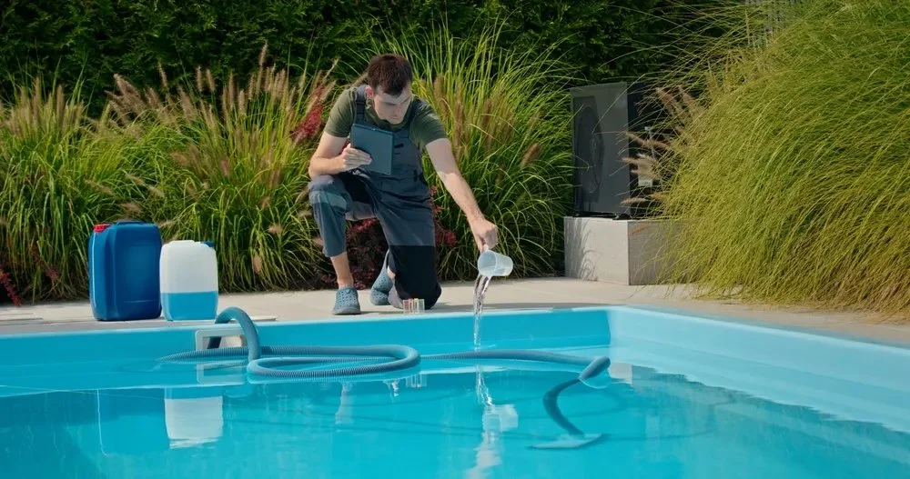 Person filling a swimming pool with a garden hose, surrounded by plants and outdoor equipment.